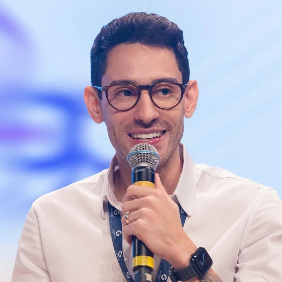 Kaio Barbosa falando ao microfone num evento, de camisa clara e óculos